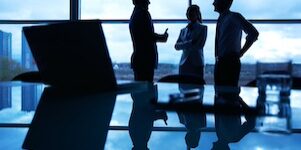 Three office workers interacting by the window with their workplace in front